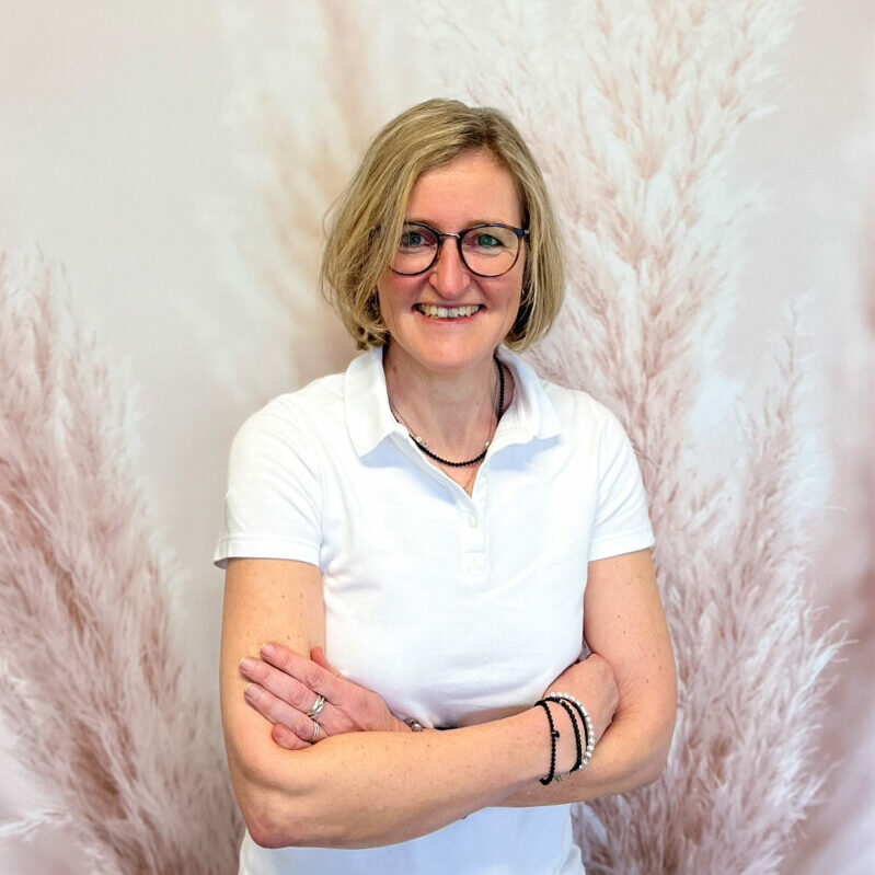 Rezeptionistin Sabine steht mit verschränkten Armen und lächelndem Blick vor einer Fotowand am Praxisstandort Kerpen-Balkhausen